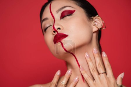 seductive asian woman with ear cuff and creative makeup posing with closed eyes and hands near neck isolated on red