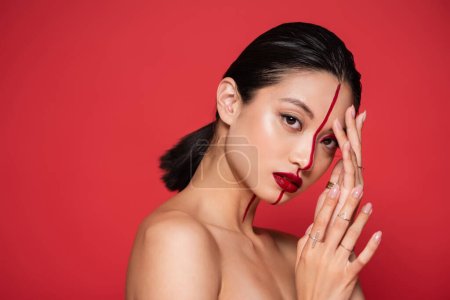 sexy mujer asiática con hombros desnudos tocando la cara con maquillaje artístico y mirando a la cámara aislada en rojo