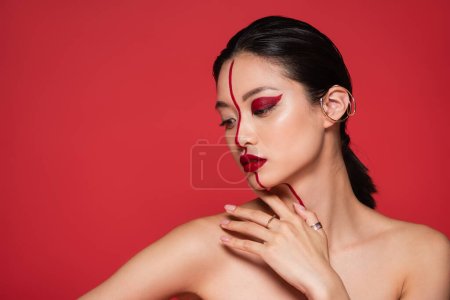 brunette asian woman with creative makeup and ear cuff posing with bare shoulders isolated on red