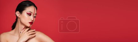 pretty and young asian woman with artistic visage touching naked shoulder isolated on red, banner