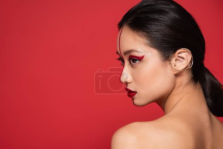 Foto de Perfil de sensual mujer asiática con maquillaje creativo mirando hacia otro lado aislado en rojo - Imagen libre de derechos