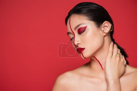 seductive asian woman with bare shoulders and artistic makeup posing with closed eyes and hand on neck isolated on red