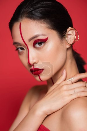 portrait of seductive asian woman with ear cuff and bright artistic visage touching bare shoulder isolated on red