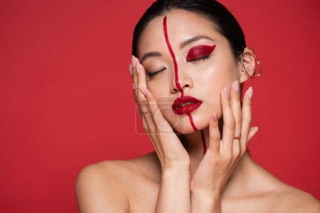 seductive asian woman with bare shoulders and artistic visage touching face and posing with closed eyes isolated on red