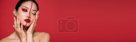 Foto de Seductora mujer asiática con hombros desnudos tocando la cara perfecta con maquillaje creativo aislado en rojo, bandera - Imagen libre de derechos