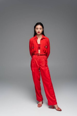 Foto de Longitud completa de la mujer asiática de moda en chaqueta roja y pantalones posando con las manos detrás sobre fondo gris - Imagen libre de derechos