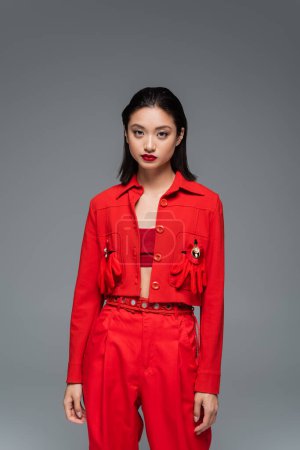 Téléchargez les photos : Jeune femme asiatique en veste rouge décorée de broches et de gants regardant la caméra isolée sur gris - en image libre de droit