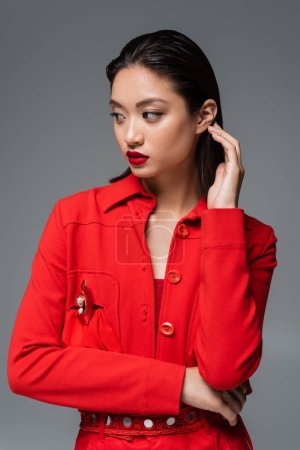 fashionable asian woman in red blazer touching brunette hair and looking away isolated on grey