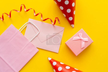 Draufsicht auf rosa Umschlag und Einkaufstasche in der Nähe von Partymützen und Geschenk auf gelbem Hintergrund 