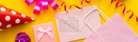 Top view of festive party cap near gift and envelope on yellow background, banner 