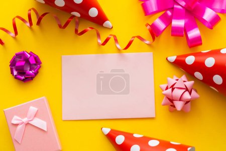 Photo for Top view of greeting card near party caps and gift box on yellow background - Royalty Free Image