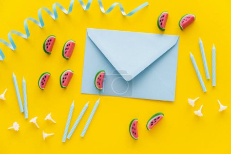 Top view of envelope near candies and festive candles on yellow background 