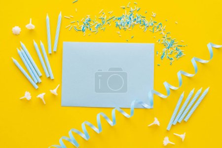 Top view of empty greeting card near candles and blue serpentine on yellow background 
