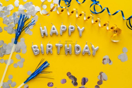 Photo for Top view of candles in shape of Happy Birthday lettering near confetti and serpentine on yellow background - Royalty Free Image