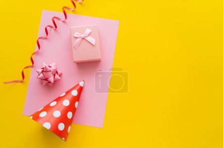 Top view of gift box on greeting card near party cap and serpentine on yellow background 
