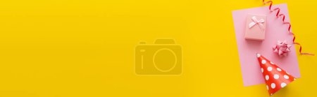 Photo for Top view of serpentine near party cap and gift box on greeting card on yellow background, banner - Royalty Free Image