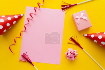 Top view of empty greeting card near present and party caps on yellow background 