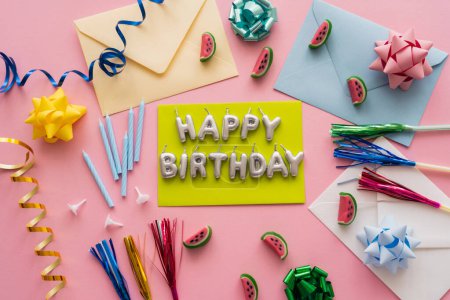 Vista superior de las velas en forma de letras de Feliz Cumpleaños cerca de sobres y caramelos sobre fondo rosa 