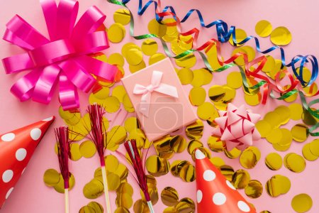 Vista superior del presente en confeti cerca de gorras de fiesta y serpentina sobre fondo rosa 