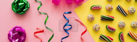 Vue du dessus des bonbons près de noeuds cadeaux colorés et serpentine sur fond rose, bannière 