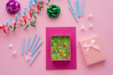 Top view of sprinkles in gift box near candles and serpentine on pink background 