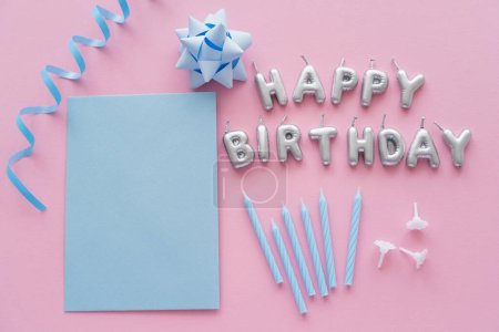 Draufsicht auf leere Grußkarte und Kerzen in Form von Happy Birthday Schriftzug in der Nähe von Geschenkschleife auf rosa Hintergrund 
