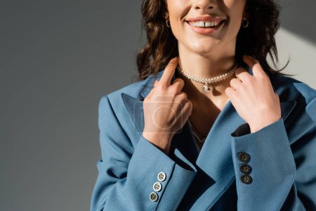 cropped view of smiling woman in pearl necklace and blue blazer on grey background