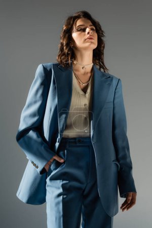 Selbstbewusste Frau im trendigen Blazer steht mit der Hand in der Tasche einer blauen Hose isoliert auf grau