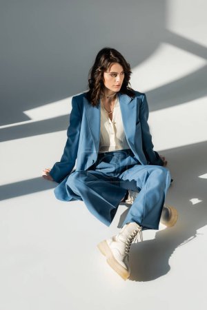 Téléchargez les photos : Pleine longueur de femme à la mode en costume bleu et bottes blanches assises sur fond gris avec éclairage et ombres - en image libre de droit