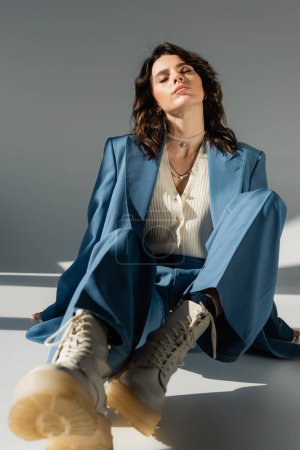 longueur totale de la femme en costume bleu et tendance et bottes lacées assis avec les yeux fermés sur fond gris