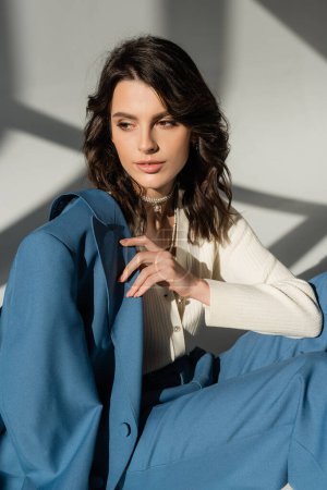 Photo for Pretty brunette woman with blue jacket looking away while sitting on grey background with lighting and shadows - Royalty Free Image