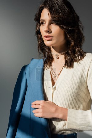 brunette woman in white cardigan and necklaces holding blue blazer and looking away isolated on grey