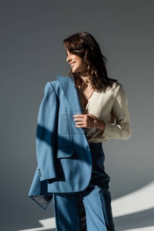 Téléchargez les photos : Femme heureuse et élégante tenant blazer bleu tout en regardant loin sur fond gris - en image libre de droit