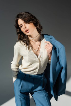 fashionable woman in white jumper and necklaces holding jacket while standing with hand in pocket of blue pants on grey background