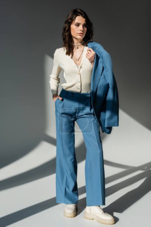 volle Länge der jungen Frau in weißem Pullover mit blauer Jacke im Stehen mit der Hand in der Tasche auf grauem Hintergrund