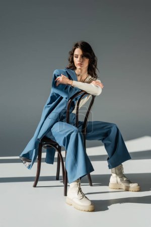 Foto de Longitud completa de la mujer de moda en pantalones y botas atadas posando en la silla cerca de chaqueta azul en gris - Imagen libre de derechos