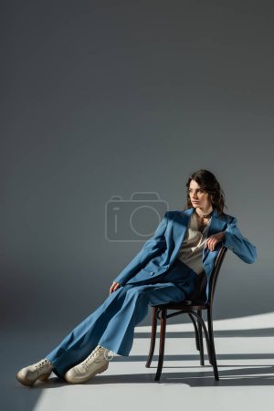 Téléchargez les photos : Longueur totale de femme élégante en costume bleu et bottes regardant loin sur la chaise sur fond gris avec éclairage et ombres - en image libre de droit