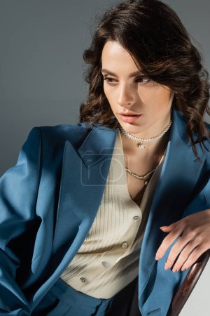 brunette woman in blue blazer and necklaces sitting and looking away on grey background