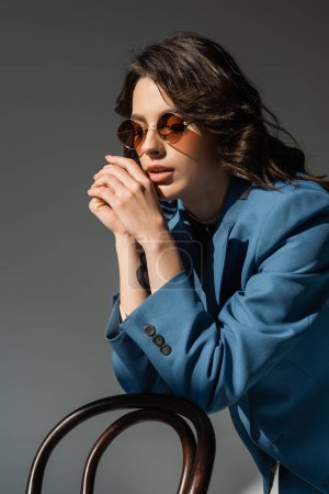 young woman in trendy sunglasses and blue jacket posing near chair isolated on grey