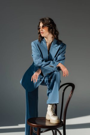 femme brune en costume bleu à la mode marchant sur la chaise et regardant loin sur fond gris
