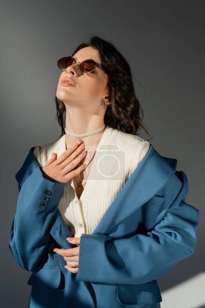 femme sensuelle en veste bleu pastel et des lunettes de soleil élégantes posant avec la main sur la poitrine sur fond gris