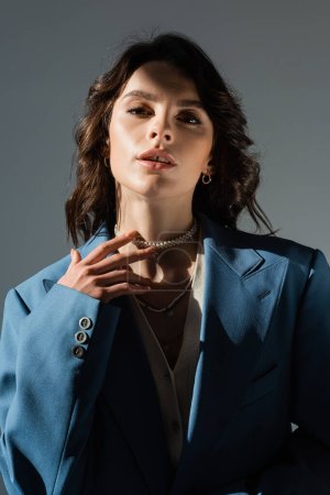 Foto de Sensual mujer morena en chaqueta azul tocando collar de perlas y mirando a la cámara aislada en gris - Imagen libre de derechos