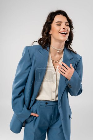 Photo for Excited woman in blue suit standing with hand in pocket and laughing with closed eyes isolated on grey - Royalty Free Image