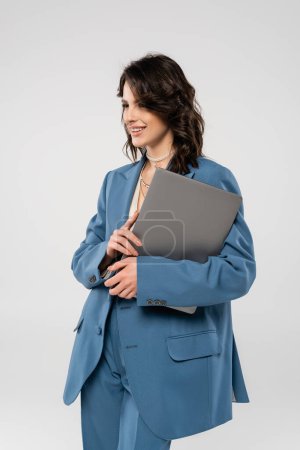 happy woman in blue blazer standing with laptop and looking away isolated on grey