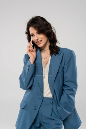 Téléchargez les photos : Femme souriante debout avec la main dans la poche de pantalon bleu et parler sur smartphone isolé sur gris - en image libre de droit