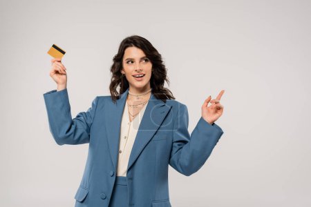 Foto de Mujer complacida en blazer azul sosteniendo tarjeta de crédito y señalando con el dedo aislado en gris - Imagen libre de derechos