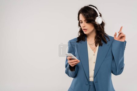 Foto de Mujer morena de moda en auriculares inalámbricos mirando el teléfono inteligente y apuntando con el dedo aislado en gris - Imagen libre de derechos