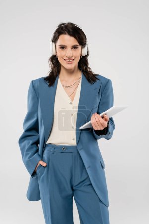 happy woman in blue suit and wireless headphones standing with hand in pocket and digital tablet isolated on grey