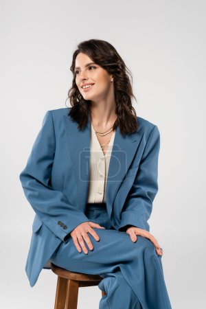 fröhliche junge Frau im blauen modischen Anzug, die auf einem Schemel sitzt und isoliert von grau wegschaut