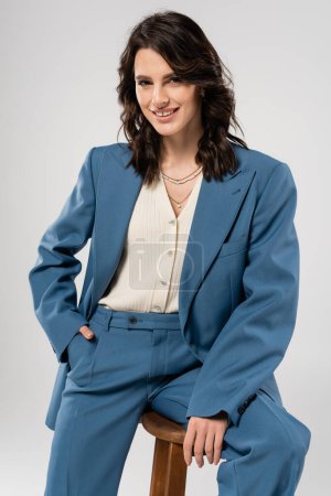 stylish brunette woman holding hand in pocket of blue pants and smiling at camera isolated on grey